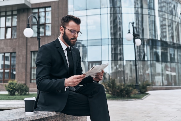 Involved with reading. Good looking young man in full suit reading a newspaper 