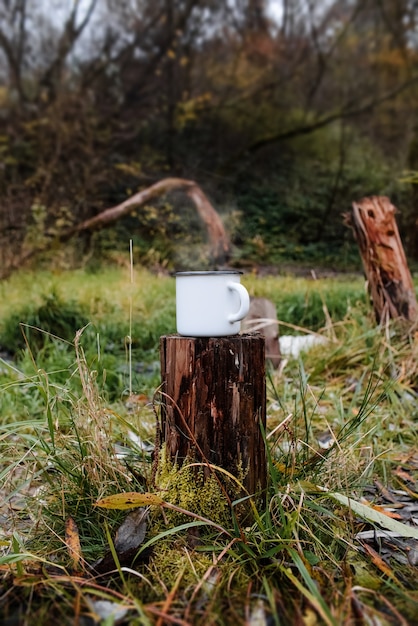 Iron mug stands on a stump in the forest. Steam rises from the hot drink.