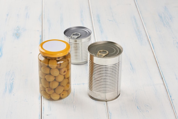 Photo iron tin can with tab opener and olives in a glass jar on the white  wooden table.