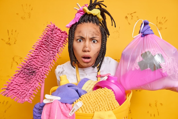 Irritated displeased woman has dirty face after cleaning fed up of housework collects rubbish in apartment holds mop stands near basket of laundry isolated over yellow wall muddy handprints.