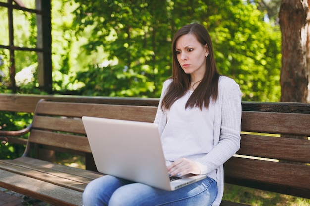 Irritated and dissatisfied businesswoman in light casual clothes. Woman sits on bench working on modern laptop pc computer in city park in outdoors on nature. Mobile Office. Freelance business concept