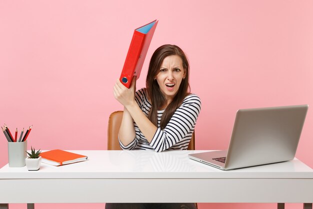 Irritated woman hiding behind red folder with paper document working on project while sit at office with laptop 