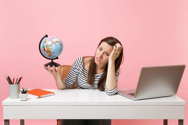 Irritated woman leaning on hand holding globe having problems with vacation planning while sit, work at office with laptop