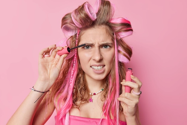 Irritated woman with long hair does hairstyle applies mascara on her long eyelashes looks angrily being in hurry for date wants to look beautiful isolated over pink background Appearance and women