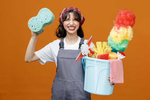 Irritated young female cleaner wearing uniform and bandana holding bucket of cleaning tools looking at camera rising sponge up isolated on orange background