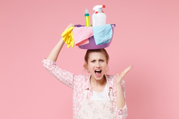 Irritated young woman housewife in apron doing housework isolated on pink wall background. Housekeeping concept. Hold basin with detergent bottles washing cleansers on head, spreading hand, screaming.