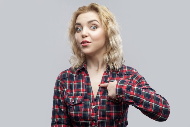 Is it me? Portrait of surprised beautiful blonde young woman in casual red checkered shirt standing, pointing herself with amazed face and asking. indoor studio shot, isolated on grey background