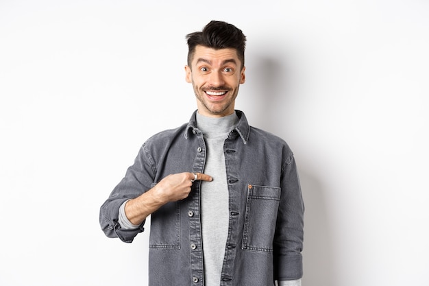 Is it me. Surprised happy guy pointing at himself, smiling cheerful, being chosen, standing on white background in spring outfit.