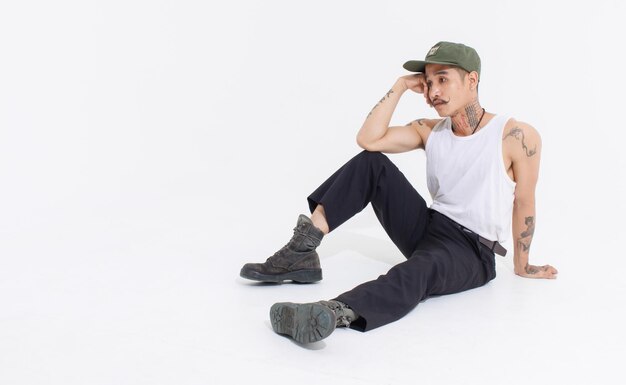 Isolated cutout studio shot Asian vintage classy mustache with neck arms hands tattoos male fashion model in casual fashionable sleeveless shirt cap boots sitting on floor on white background