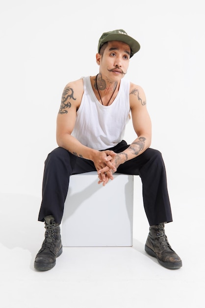 Isolated cutout studio shot Asian vintage classy mustache with neck arms hands tattoos male fashion model in casual fashionable sleeveless shirt cap boots sitting on square box on white background