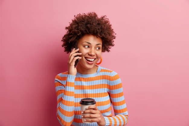 Isolated shot of good looking woman with Afro hair looks happily away keeps smartphone near ear drinks takeaway coffee wears casual striped jumper isolated over pink wall