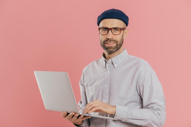 Isolated shot of handsome unshaven male does distance job on portable laptop computer wears hat and optical glasses connected to wireless internet stands over pink background develops software