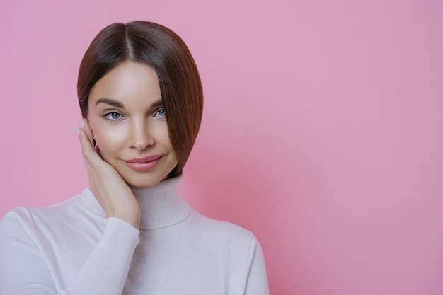 Isolated shot of pleasant looking dark haired young woman has well groomed face dressed in turtleneck jumper has healthy skin isolated over rosy background People beauty wellbeing concept