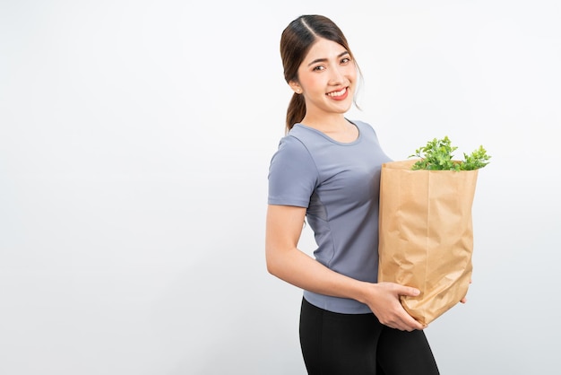 Isolated a young healthy attractive Asian woman smile and wears sporty clothes carrying vegetables in a grocery paper bag with white background