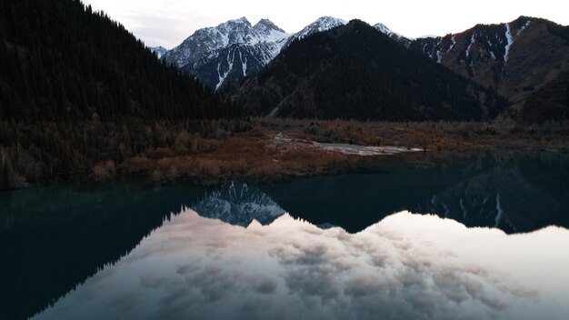 Issyk mountain lake with mirror water at sunset. Snowy mountains, forest and clouds.