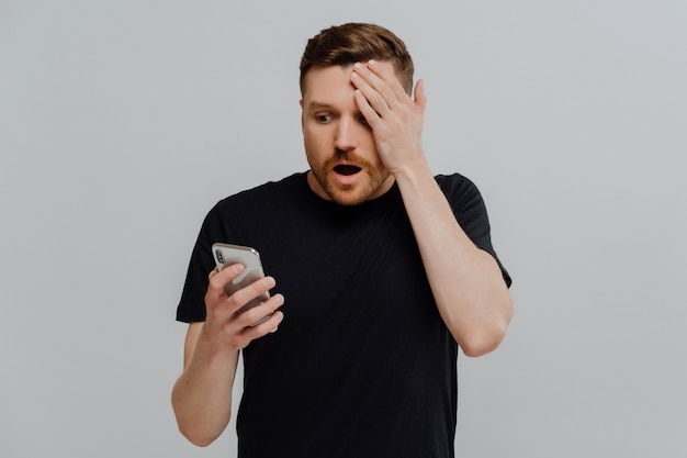 It can not be true. Young impressed unshaved ginger man reading latest news in internet on mobile phone, being in shock while receiving shocking message on smartphone, isolated over grey background