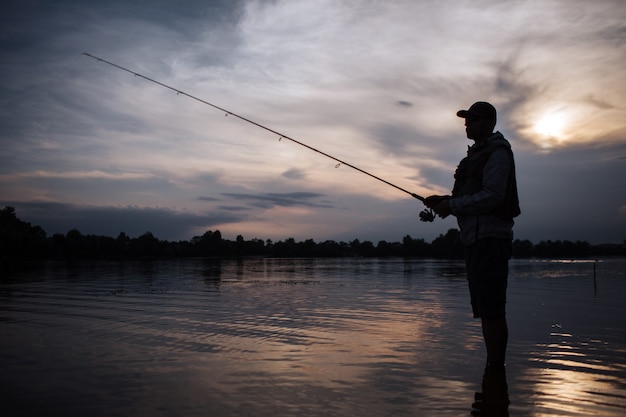 It is evening outside. Fisherman stands in shallow and fishing. He holds fly rod in hands. There is a reel under it. He stands without moving.