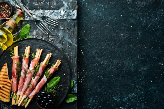 Italian antipasto Bread sticks grissini with prosciutto ham on a black plate Rustic style Top view Free space for your text