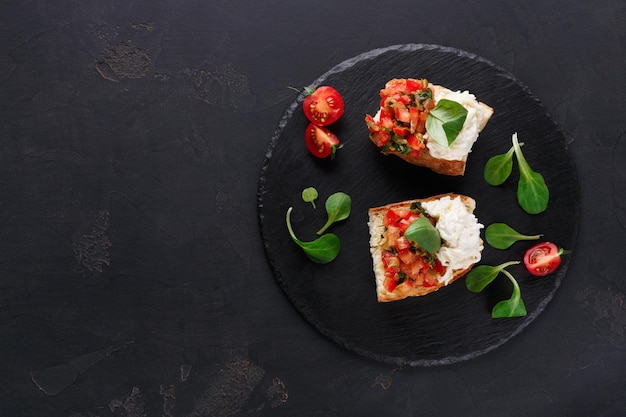 Italian appetizer on black background. Crusty bruschetta with concasse tomatoes, stracciatella cheese decorated with spinach. Delicious and healthy restaurant meals, top view, copy space