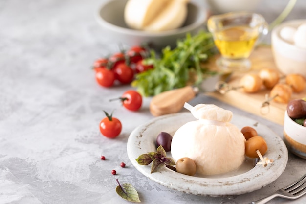 Italian cheeses burrata, mozzarella, scorocia, boccini with olives, cherry tomatoes and arugula on a round white plate on a light background. Copy space.