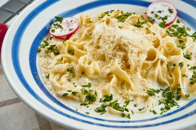 Photo italian fettuccine pasta in cheese sauce with parmesan, cherry tomatoes and herbs in white ceramic bowl on white surface.