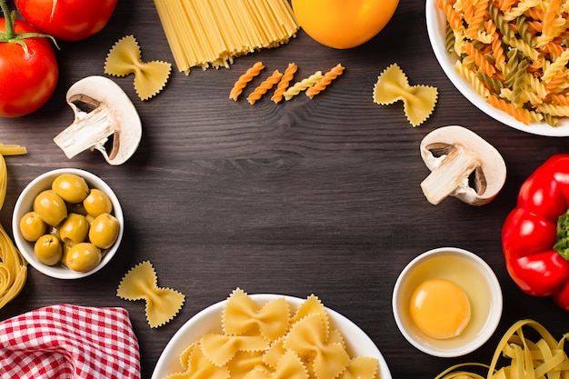 Italian food ingredients frame with various pasta, vegetables, mushrooms, olives. Flat lay on dark wooden background