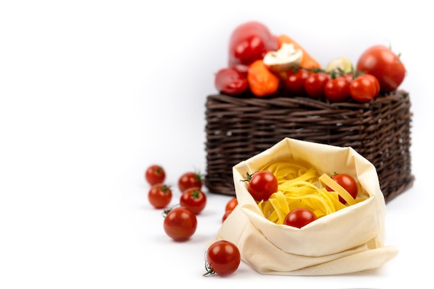 Italian pasta Tagliatelle or Fettuccine in a cloth bag with cherry tomatoes and a wicker basket with vegetables on a white background side view Copy space