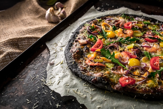 Photo italian pizza with black dough and seafood on a baking tray from the oven. on top of it lay squid, shrimp, octopus, onions, tomatoes. cuttlefish ink