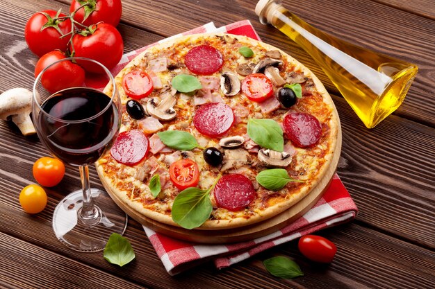 Italian pizza with pepperoni, tomatoes, olives, basil and red wine on wooden table. Top view