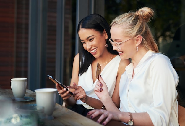 Its really that easy Shot of a woman using a cellphone while her friend holds up a credit card while sitting together