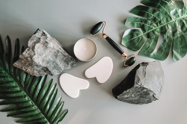 Jade facial roller and gua sha scraper and natural stone with greenery on a gray background Facial massage set for lifting therapy made of natural stones Flat lay top view