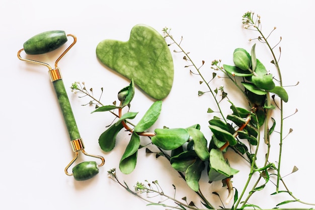 Jade roller massager and gouache facial scraper on a background with green plants