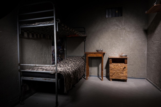 Jail cell with iron bunk bed and wooden bedside table with aluminum dishes