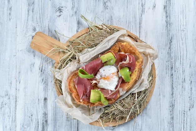 Jamon on French toast with poached egg and avocado on a wooden board and straw. top view