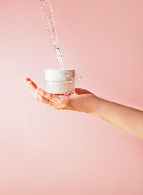 Jar of cream in a female hand with pouring water and splashes on a pink background