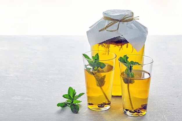 Jar of kombucha and two glass of kombucha are garnished with mint and lime lemon on a light gray concrete table