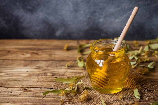 Photo a jar of liquid honey from linden flowers and a stick with honey on a dark surface. copy space