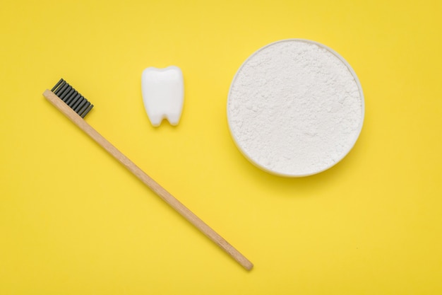 A jar of tooth cleaning powder, a wooden toothbrush and a tooth figurine on a yellow table.The view from the top. Flat lay.