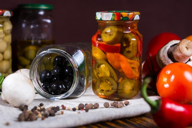 Jar with black olives lies on a napkin near jars of pickled vegetable, spices, tomatoes, mushrooms on wooden table