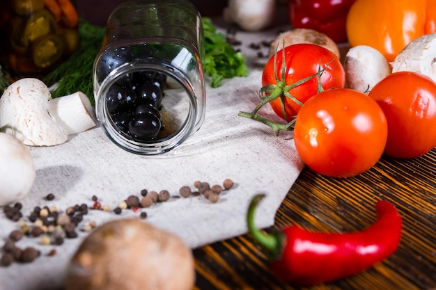 Jar with black olives lies on a napkin near spices, tomatoes, mushrooms and other vegetables on wooden table