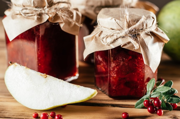 Jars of homemade lingonberry and pear jam with craft paper on lids on wooden table next to fresh lingonberries and pears