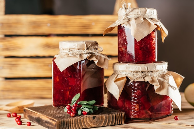 Jars of homemade lingonberry and pear jam with craft paper on lids on wooden table, next to fresh lingonberries