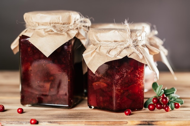 Jars of homemade lingonberry and pear jam with craft paper on lids on wooden table next to fresh lingonberries