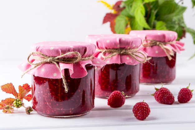 Jars with raspberry jam placed in rows and fresh raspberry on white wooden table