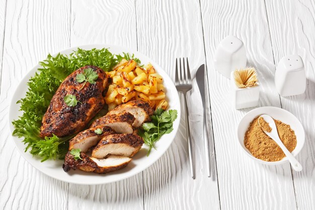 Jerk chicken breasts with grilled ananas cubes and fresh green leaves lettuce on a white plate on a wooden textured table horizontal view from above