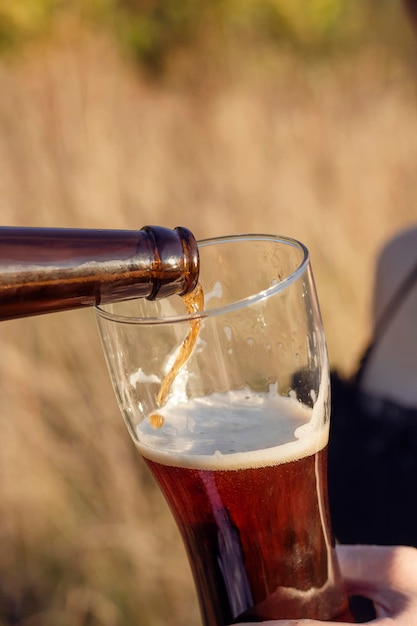 A jet of dark beer from a bottle pours into a glass glass in the hands of a girl