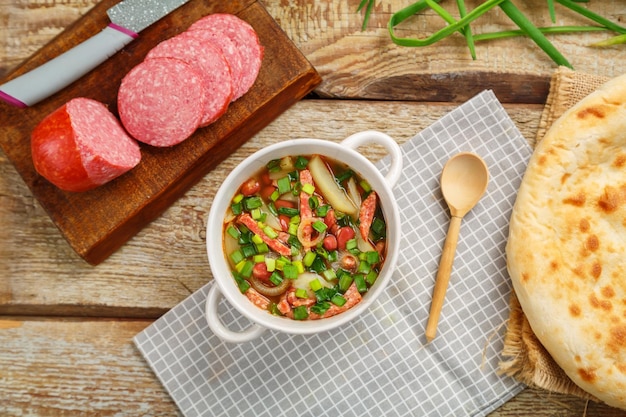 Jewish bean soup with smoked meats in a tureen on a gray napkin next to a sausage and a flatbread