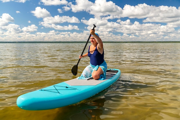 A Jewish woman in a kerchief kisuy rosh on her head and in a pareo on her knees on a SUP board with an oar swims in the lake on a sunny day