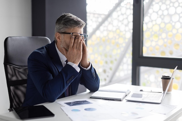 Job Burnout Exhausted Mature Businessman Sitting At Workplace And Rubbing Eyes