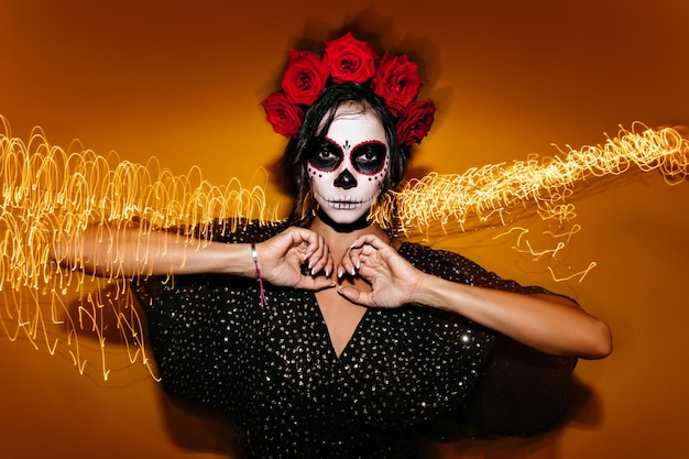 Jocund tanned woman with zombie face painting posing in studio Cheerful brunette girl in red wreath celebrating halloween